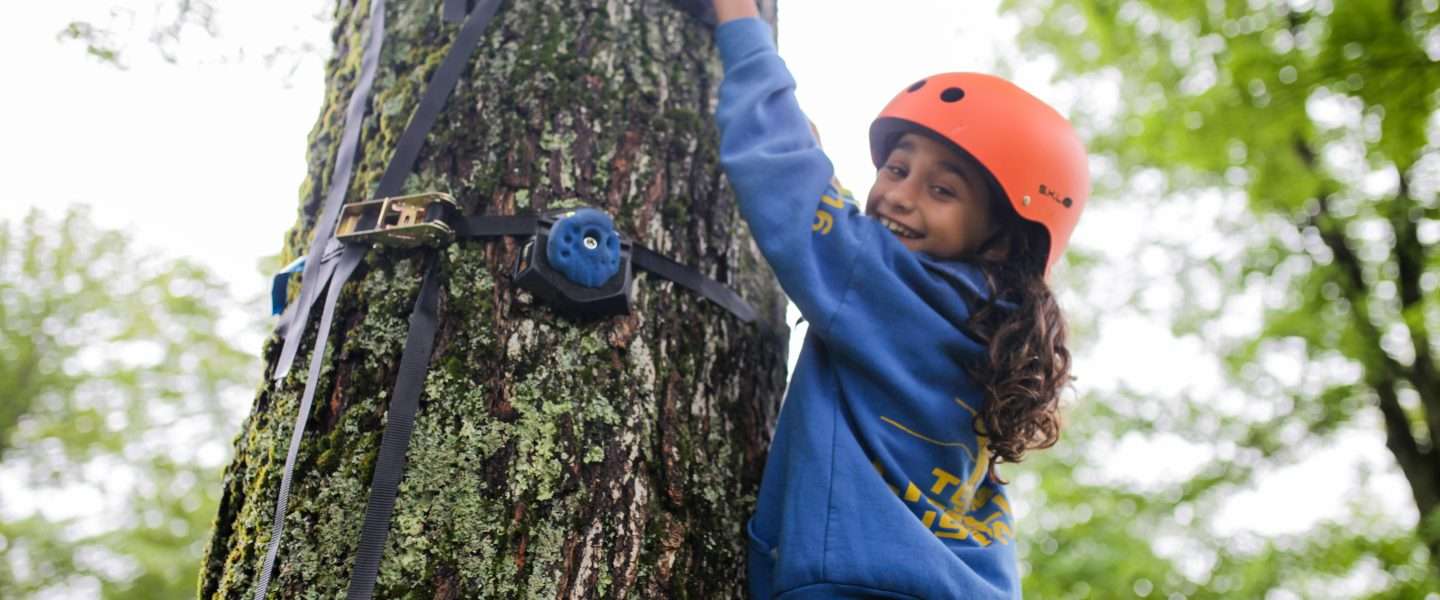 Female camper in a blue top climbing a tree with full safety gear