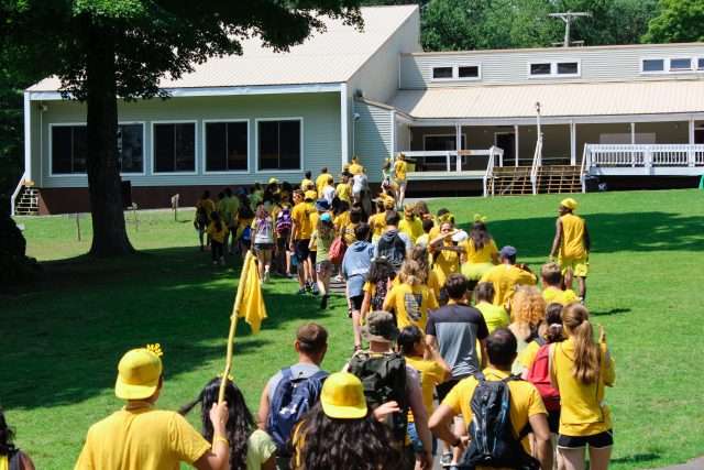 Campers being led to their dorms by staff, one long line snaking through the green grass surrounding the bunk buildings